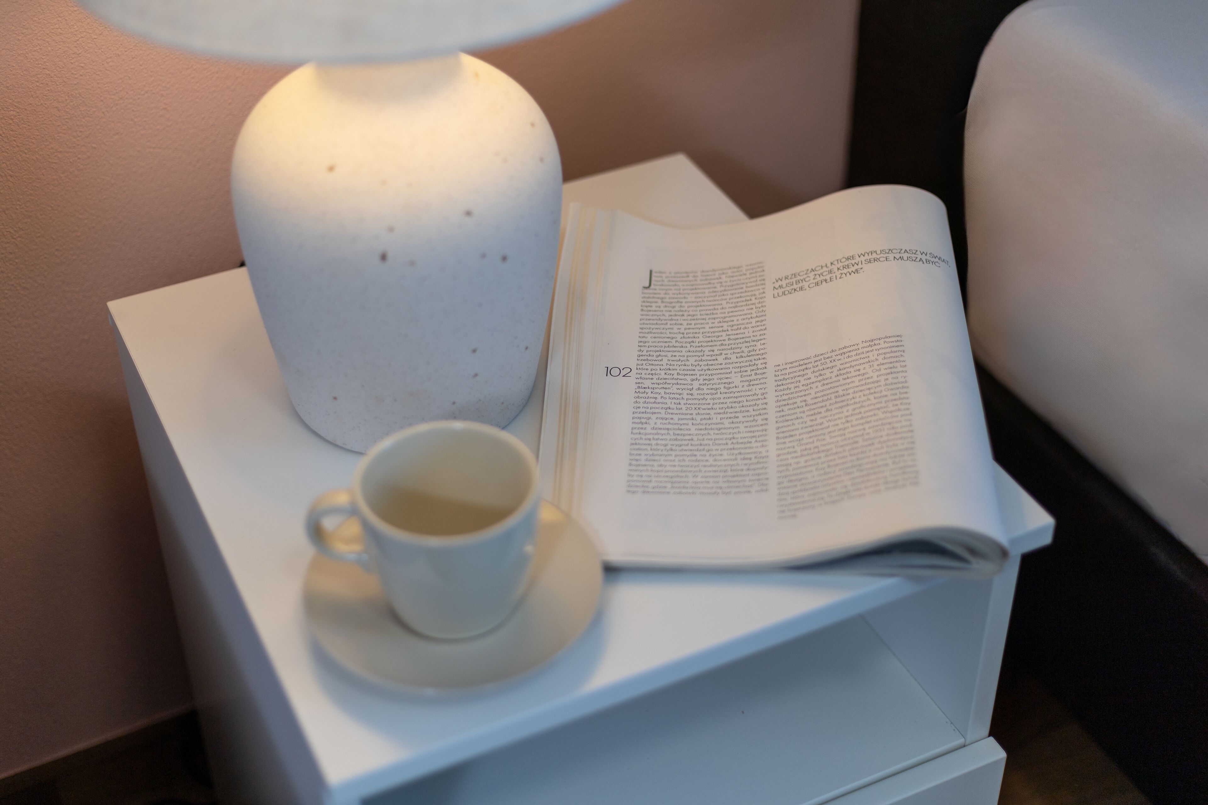 A white bedside table with a lamp, a cup of tea, and an open book.