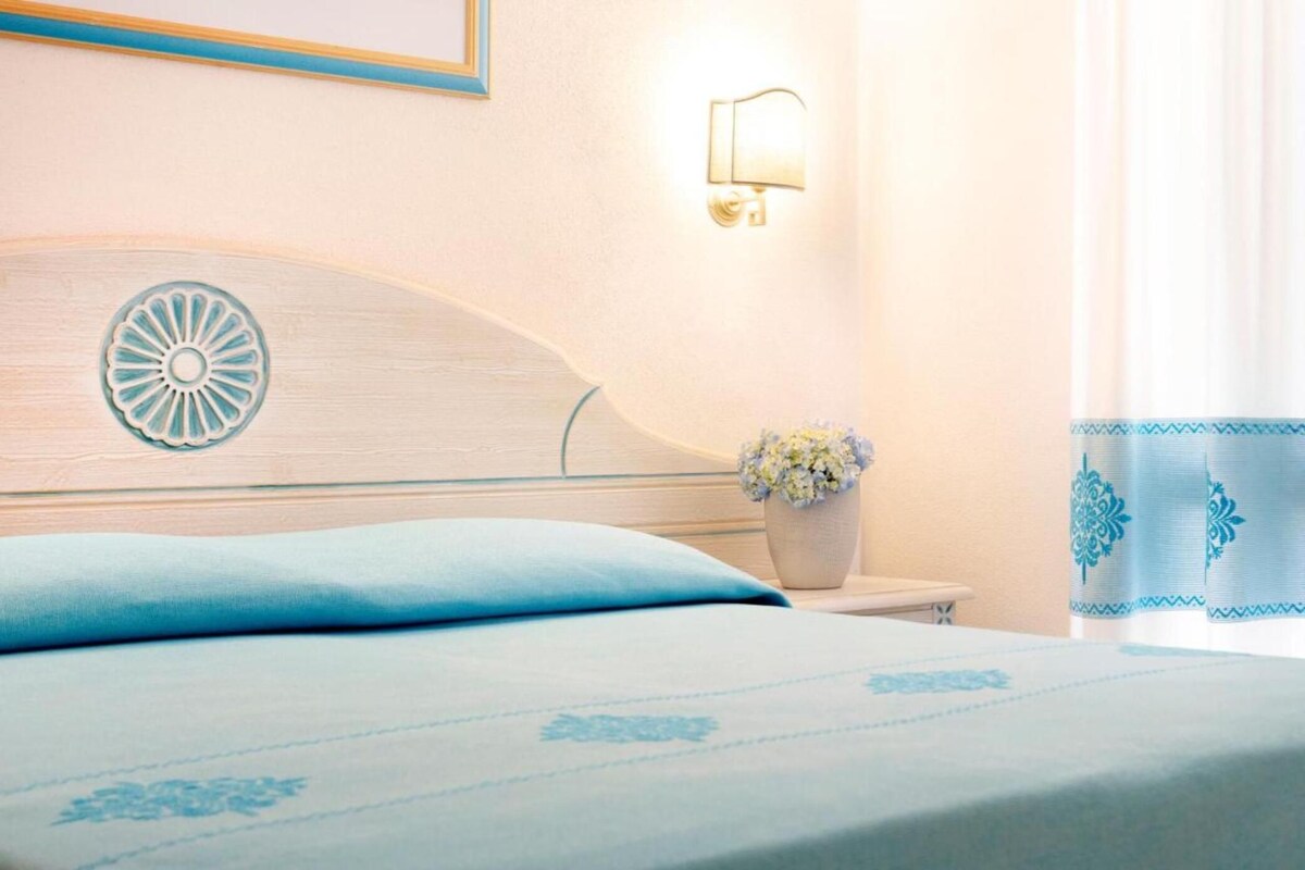 Una cama bien hecha con una colcha azul claro y almohadas, un cabecero decorativo con un diseño circular, una mesita de noche con un jarrón de flores y una lámpara de pared.
