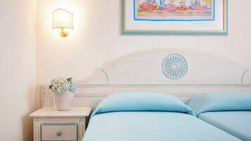 A neatly made bed with light blue bedding, a bedside table with a vase of flowers, a wall-mounted lamp, and a framed painting above the bed.