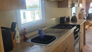 A compact kitchen with a gas stove, a sink, and a window with blinds. - 3 Bed Caravan - Sleeps 6 - Pets - Parking - Wifi (Borth)