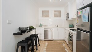 A modern kitchen with white cabinetry, a dishwasher, a stove, and a refrigerator. There are two bar stools and a small plant on the counter. - Hidden Gem - 150m from Urunga Sea Lido (Urunga)