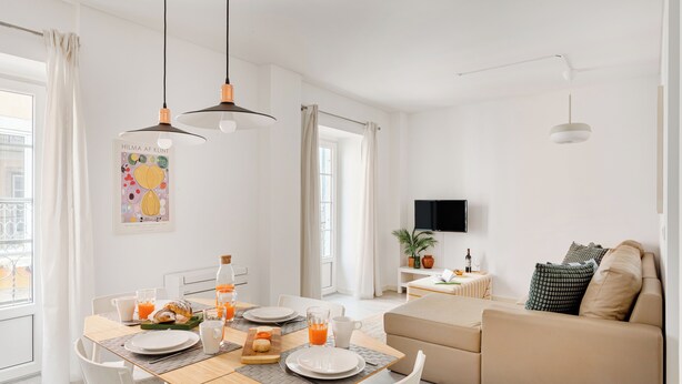 Una sala de estar moderna y bien iluminada, con una mesa de comedor de madera dispuesta para el desayuno, un sofá y un televisor montado en la pared.