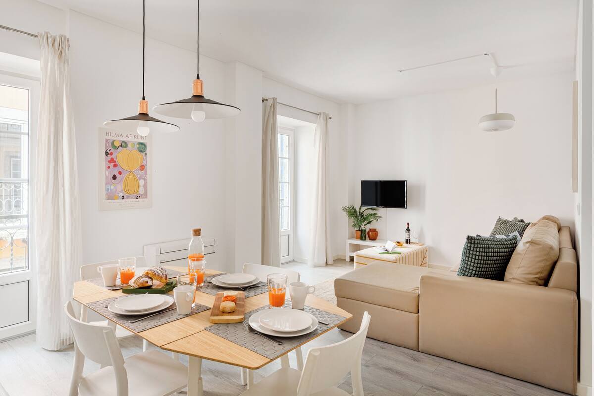 A modern, well-lit living room with a wooden dining table set for breakfast, a sofa, and a TV mounted on the wall.