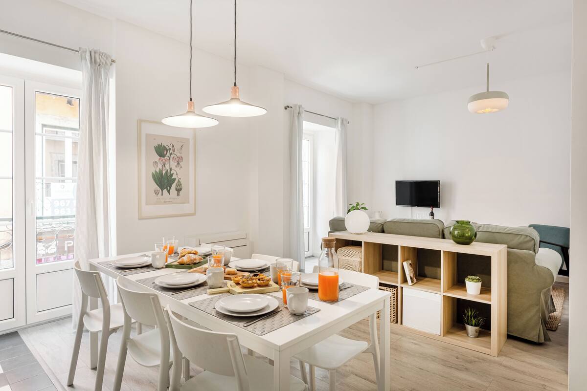 A modern dining area with a white table set for breakfast, a sofa, and a TV in the background.
