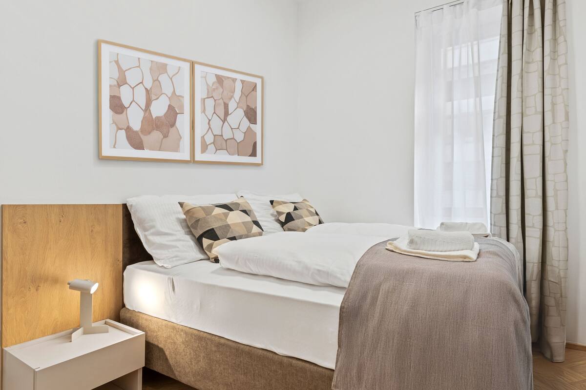 Quarto moderno com cabeceira de madeira, mesa de cabeceira com luminária, cama com roupa de cama branca e almofadas estampadas, e arte abstrata na parede.