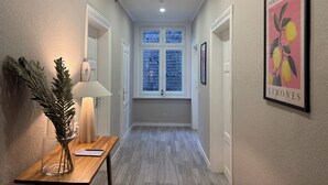 A hallway with a wooden floor, a window with blinds, a framed picture of lemons, and a vase with green foliage.