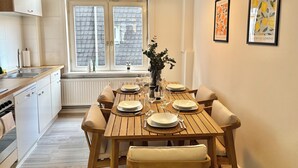 A dining area with a wooden table set for four, a window with a view of the roof, and two framed artworks on the wall.