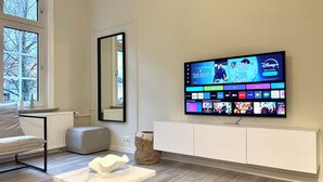A modern living room with a flat-screen TV mounted on the wall, a white sofa, a small table with a white bowl, and a large window with a view of trees.
