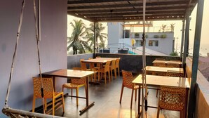 A covered outdoor seating area with tables and chairs, a hanging hammock, and a view of the sea and buildings in the background. - Small Daddy Plus (Calangute)