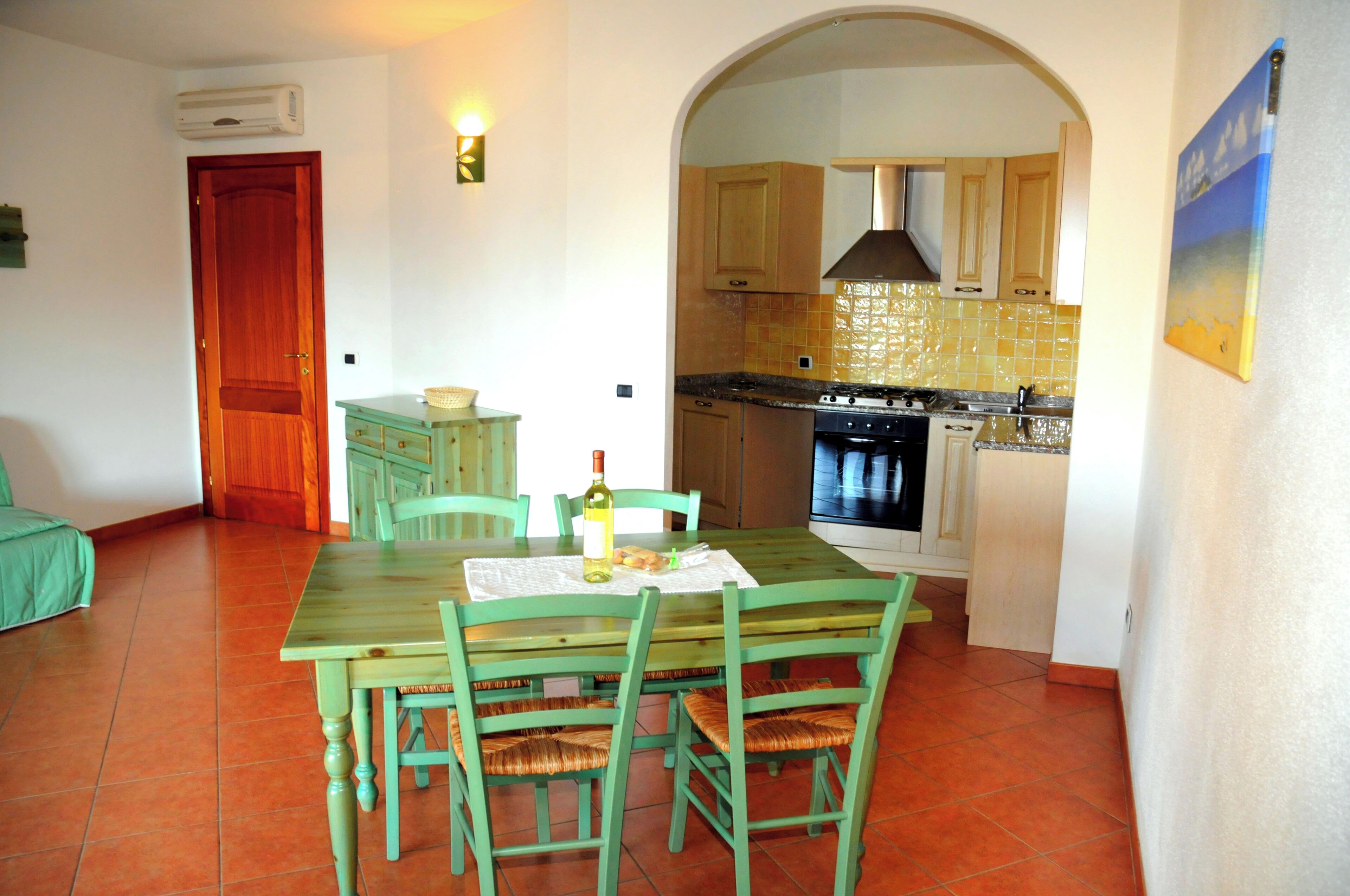 Un espace repas avec une table et des chaises en bois, une cuisine visible à travers une arche, et une porte donnant sur une autre pièce.