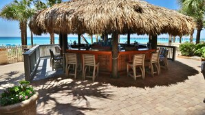A thatched-roof outdoor bar area with wooden chairs and a table, overlooking a beach with palm trees and a clear blue sky. - 3BR 2BA @ The Calypso Breathtaking View, Walk to Pier Park (Panama City Beach)
