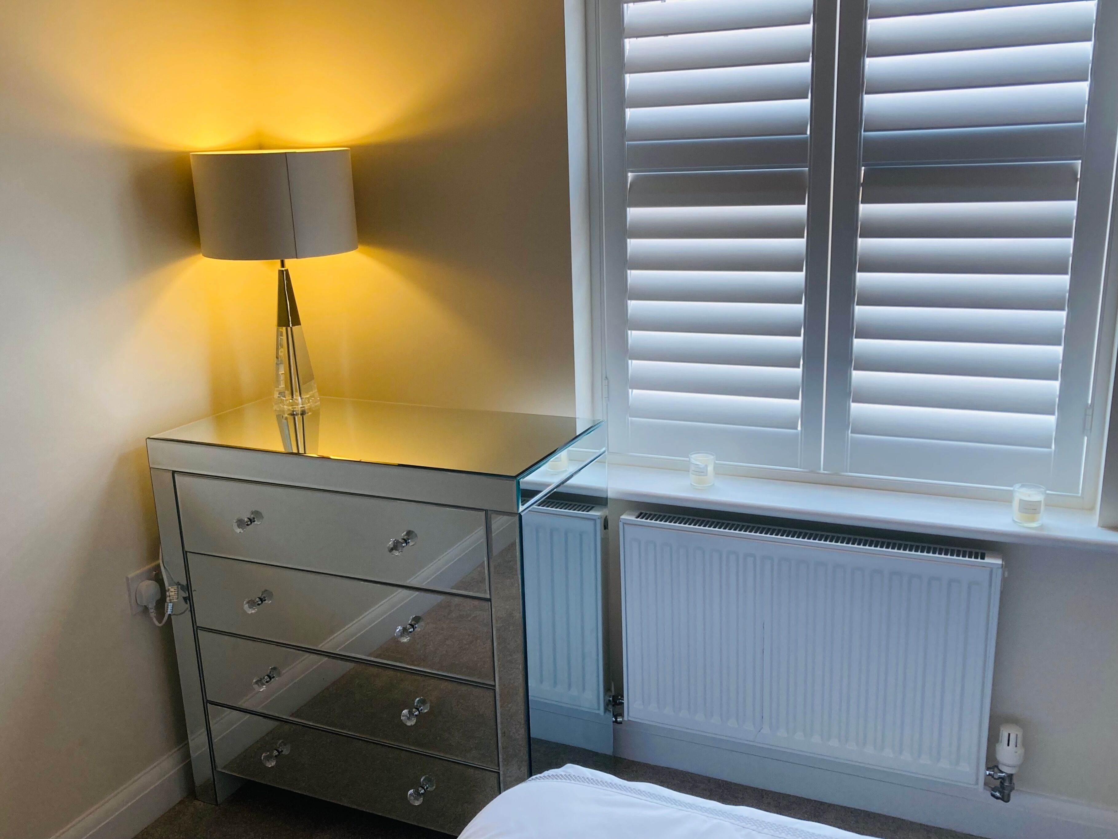 A bedroom with a glass-top dresser, a bedside lamp, a radiator, and a window with blinds.