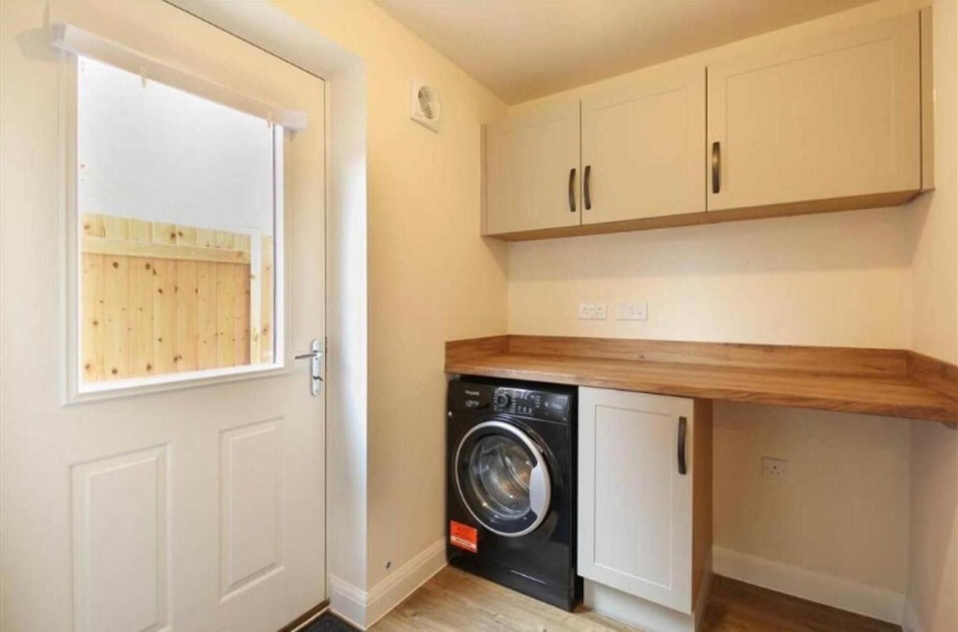 Une petite buanderie comprenant un lave-linge, des placards en bois et une porte donnant sur une clôture en bois.