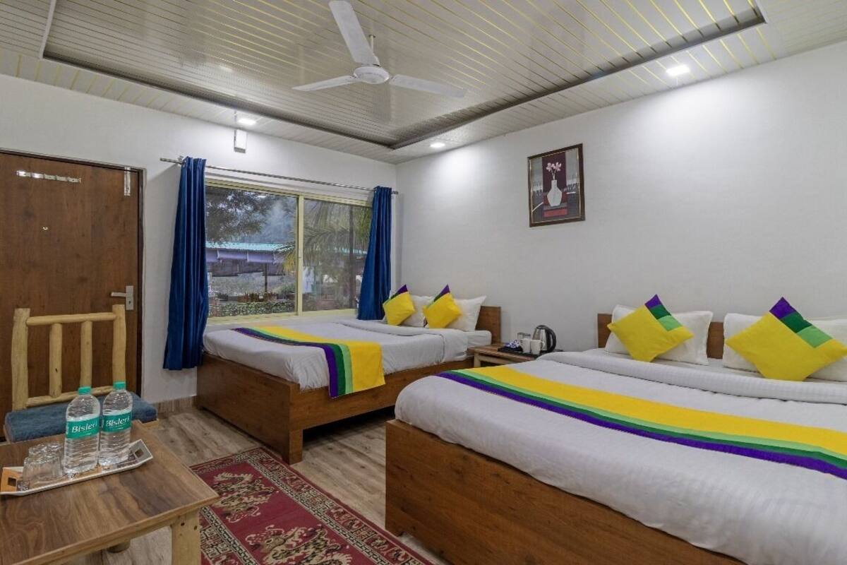 Une chambre d’hôtel avec deux lits en bois, une table en bois sur laquelle sont posées des bouteilles d’eau, un tapis rouge à motifs et un ventilateur de plafond.