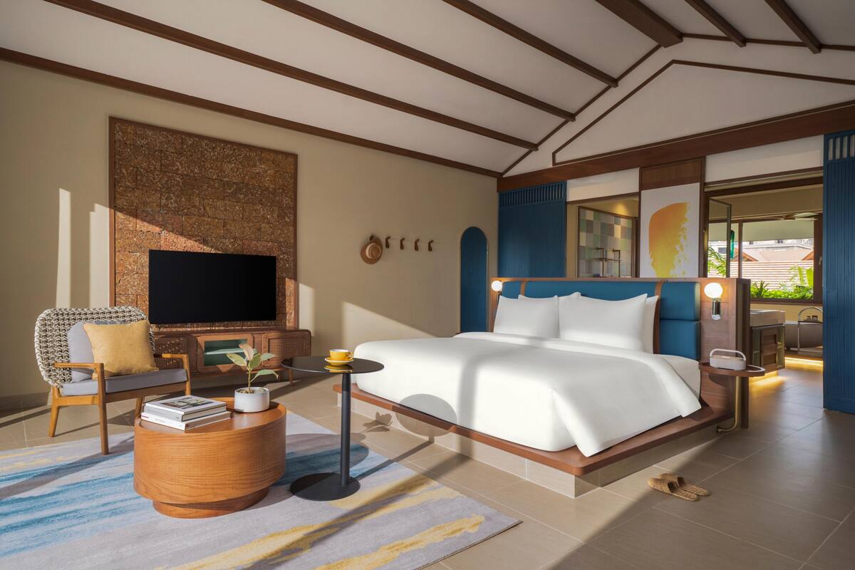Habitación de hotel con una cama grande, un televisor de pantalla plana, una zona de estar con una silla y una mesita, y vistas al exterior.