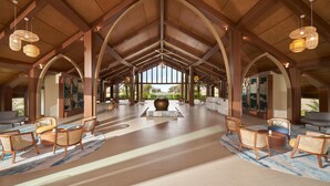 Eine großzügige Lobby mit hohen Decken, Holzbalken und einer zentralen Sitzecke mit Rattan-Stühlen und einer großen Vase.