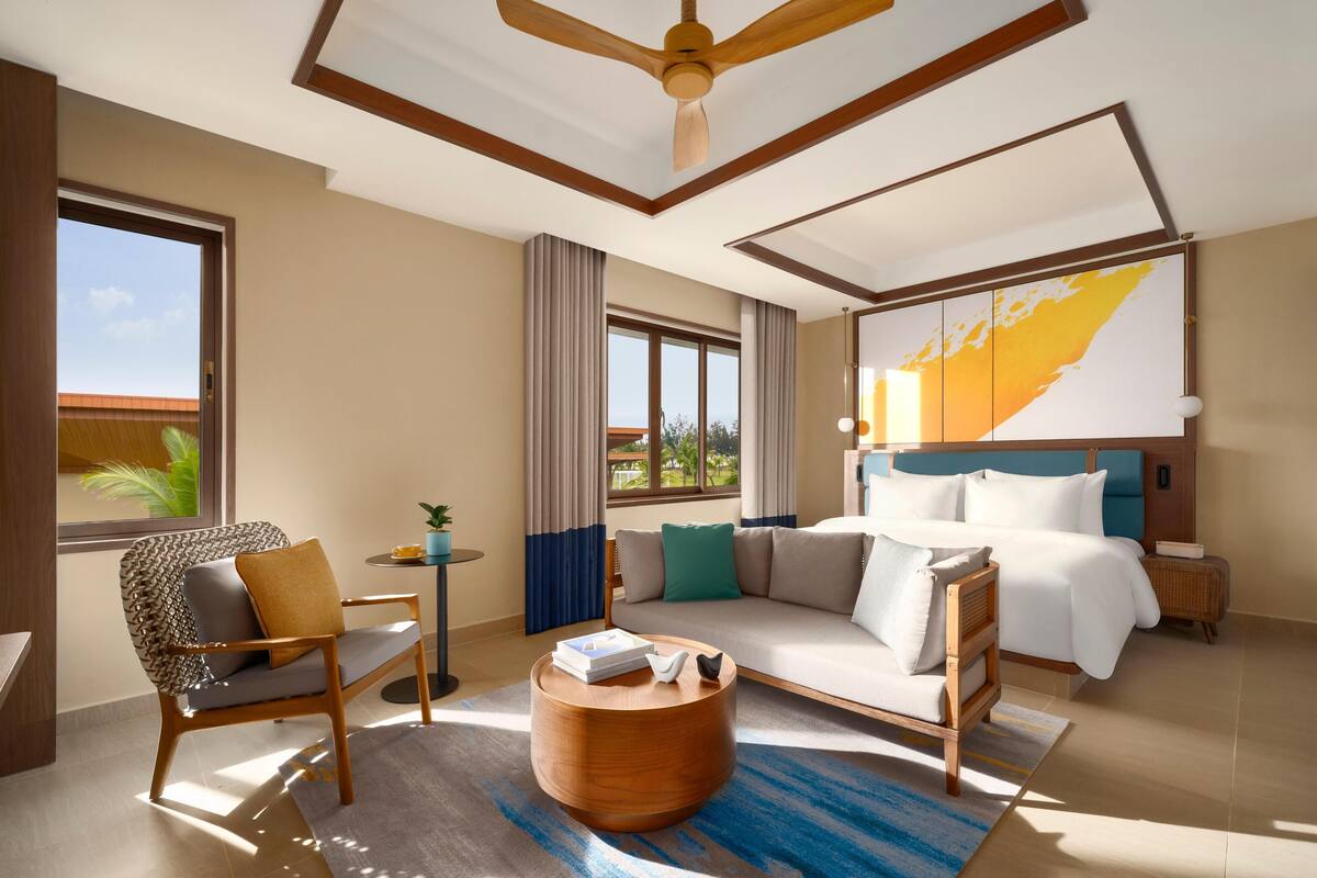 Una habitación de hotel moderna con cama, sofá, sillón, mesa de centro redonda y ventilador de techo.