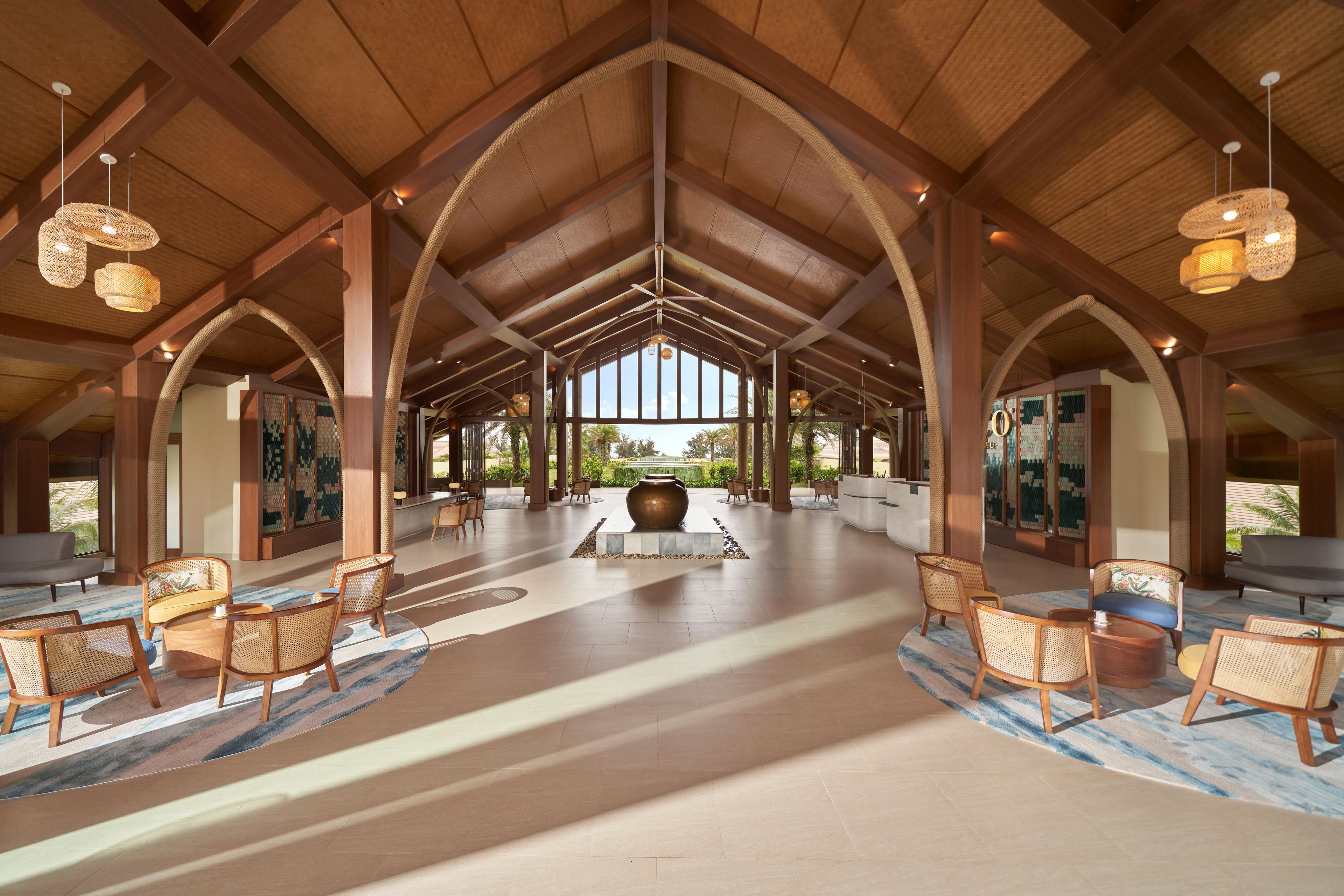 A spacious lobby with high ceilings, wooden beams, and a central seating area with rattan chairs and a large vase.
