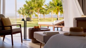 Ein Hotelzimmer mit Blick auf Palmen und den Ozean, ein Bett, ein Sessel, ein kleiner Tisch mit einer Pflanze und ein Hut auf dem Bett.