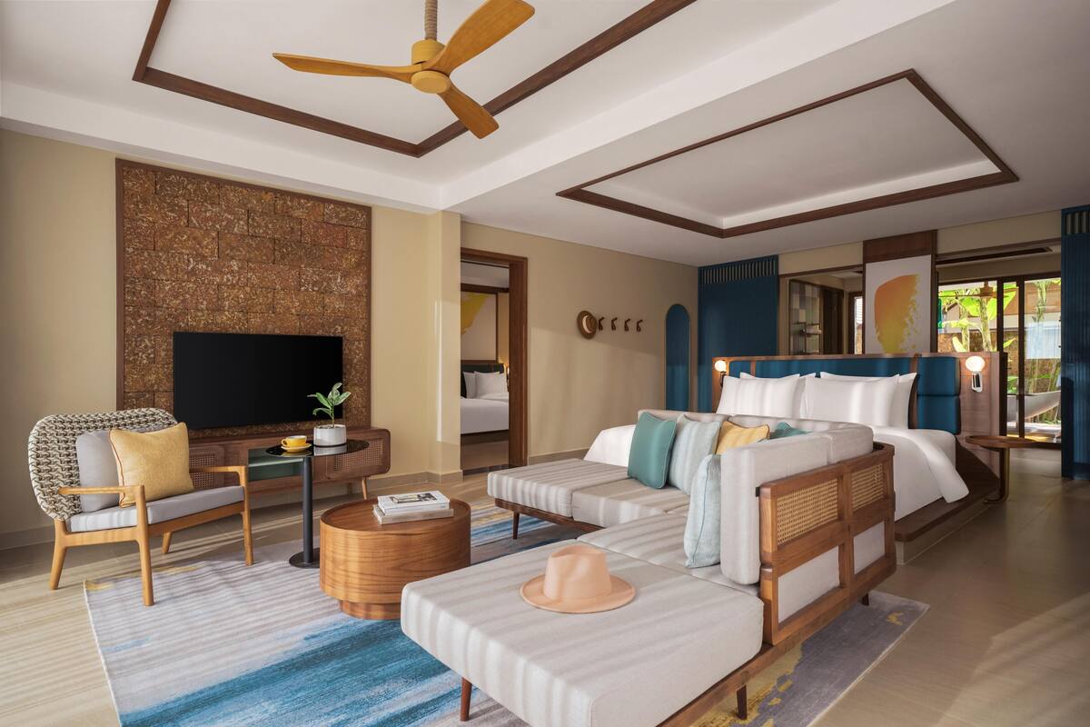 Una habitación de hotel moderna con una televisión de pantalla plana, un sofá, una cama con cortinas azules, un ventilador de techo de madera y una pared con diseño de ladrillo.