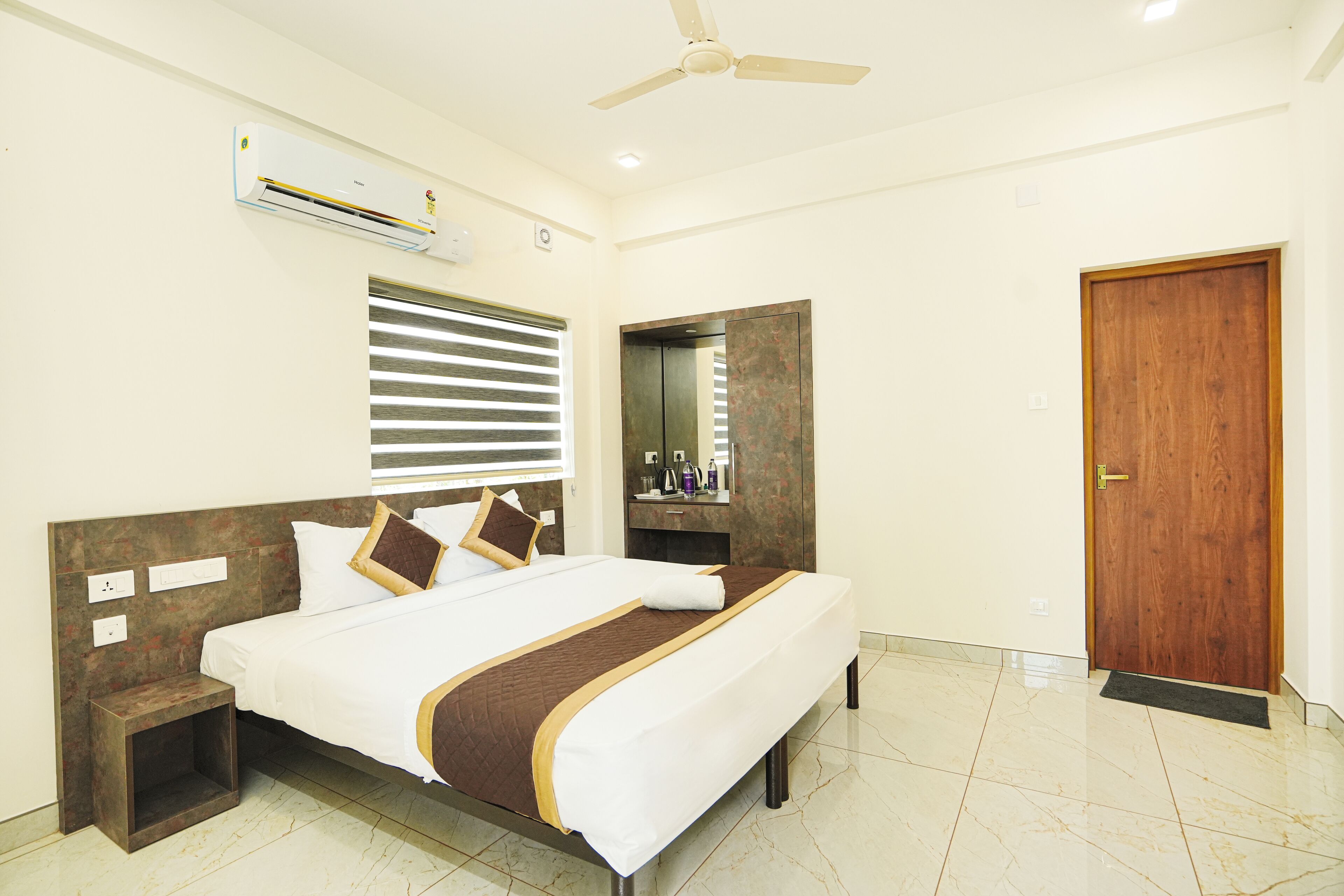Habitación de hotel con cama, mesita de noche, equipo de aire acondicionado, ventana con persianas, espejo y puerta de madera.