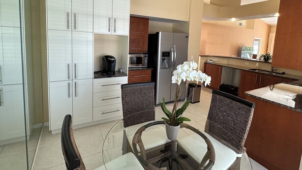 Una cocina moderna con armarios blancos, un refrigerador, un horno y una cafetera. Hay una mesa de comedor con sillas y un florero con flores blancas.