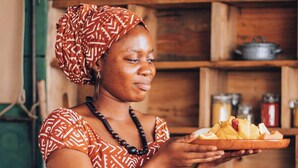A person holding a plate with food, standing in front of a wooden shelf with jars. - Rufiji Explorers (Mibuyusaba)