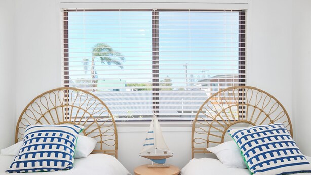 Two single beds with wicker headboards, white bedding, and patterned pillows. A small wooden table with a sailboat model is between the beds.