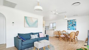 A modern living room with a blue sofa, a dining area with wicker chairs, and a kitchen in the background.