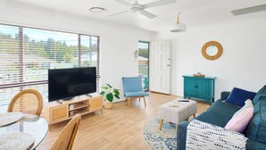 A modern living room with a flat-screen TV, wicker furniture, a teal cabinet, and a large window with blinds.