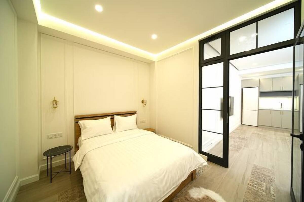 A bedroom with a bed, bedside tables, a small stool, and a view into a kitchen area through a glass door.