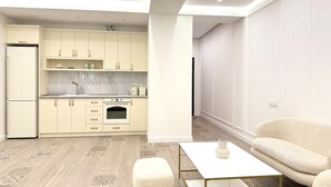 A modern kitchen with light-colored cabinets, a white countertop, and a built-in oven. There is a white sofa and a small table with a vase in the living area. - Sweet Home Prime Apart-Hotel (Yerevan)