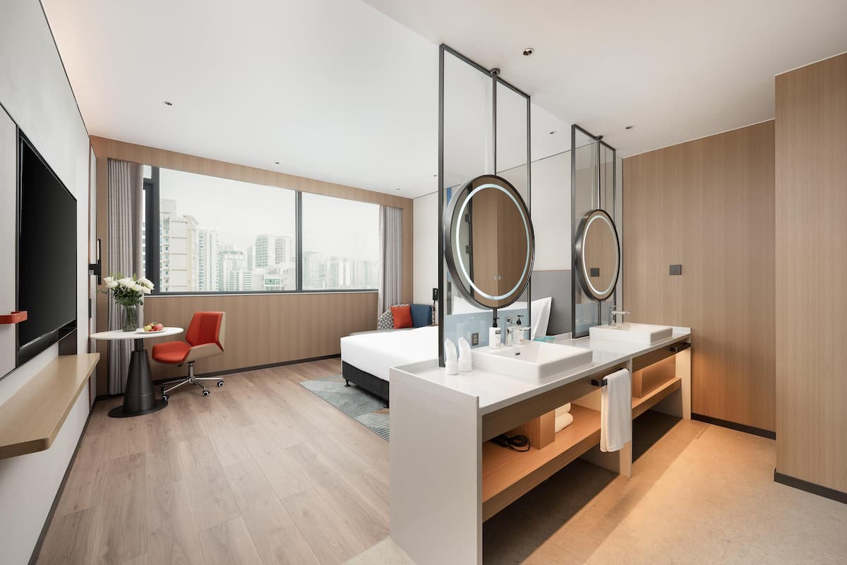 Una habitación de hotel moderna con una cama grande, un escritorio con una silla, un tocador con dos lavabos y un ventanal con vistas a la ciudad.