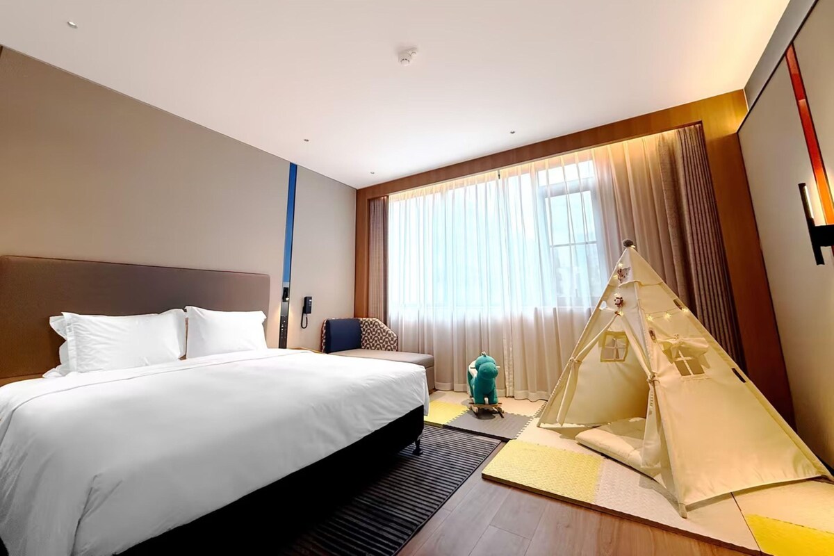 A hotel room with a large bed, a small seating area, a tent-like structure, and a large window with curtains.