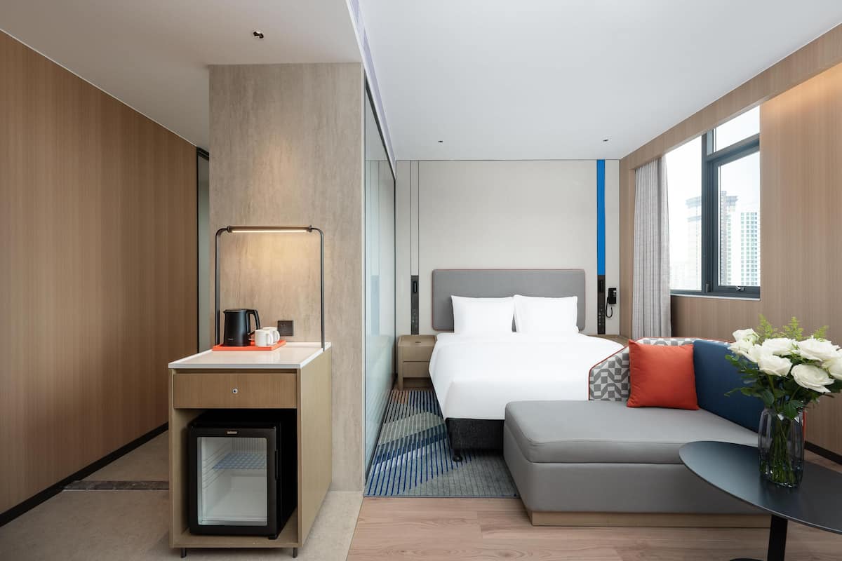 Una habitación de hotel moderna con cama, sofá, escritorio con cafetera, refrigerador pequeño y ventana con vista a la ciudad.