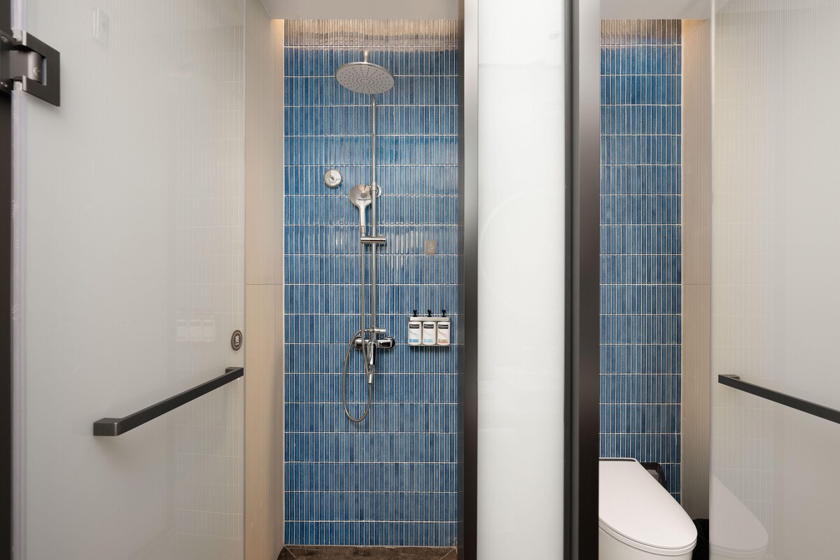 A bathroom with a blue tiled wall, a showerhead, a toilet, and a handrail.