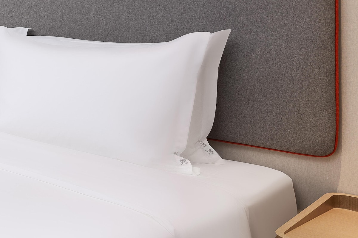 Una cama bien tendida con ropa blanca y almohadillas, cabecera gris y mesita de noche de madera.