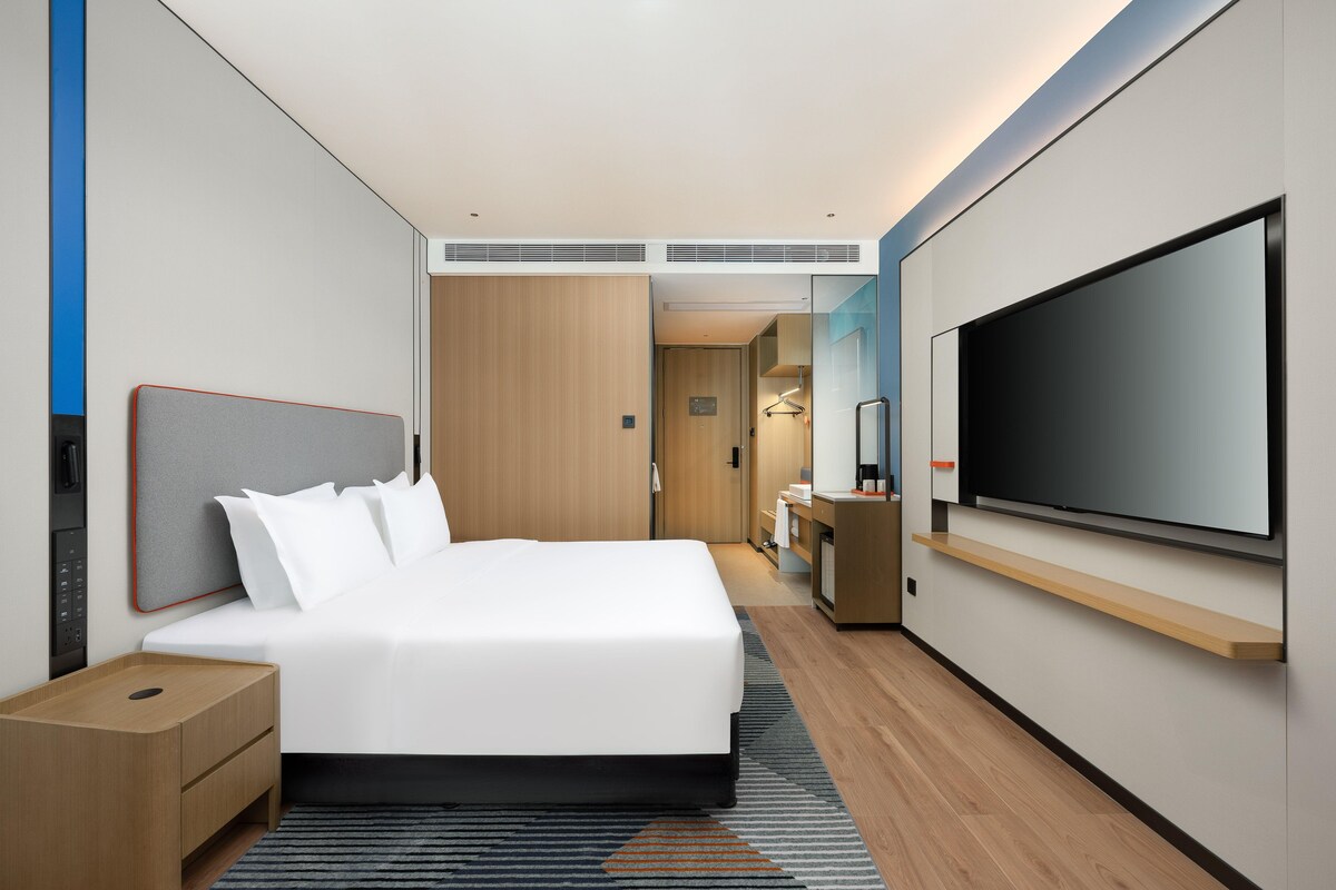 A modern hotel room with a large bed, a flat-screen TV, a wooden nightstand, and a desk with a chair.