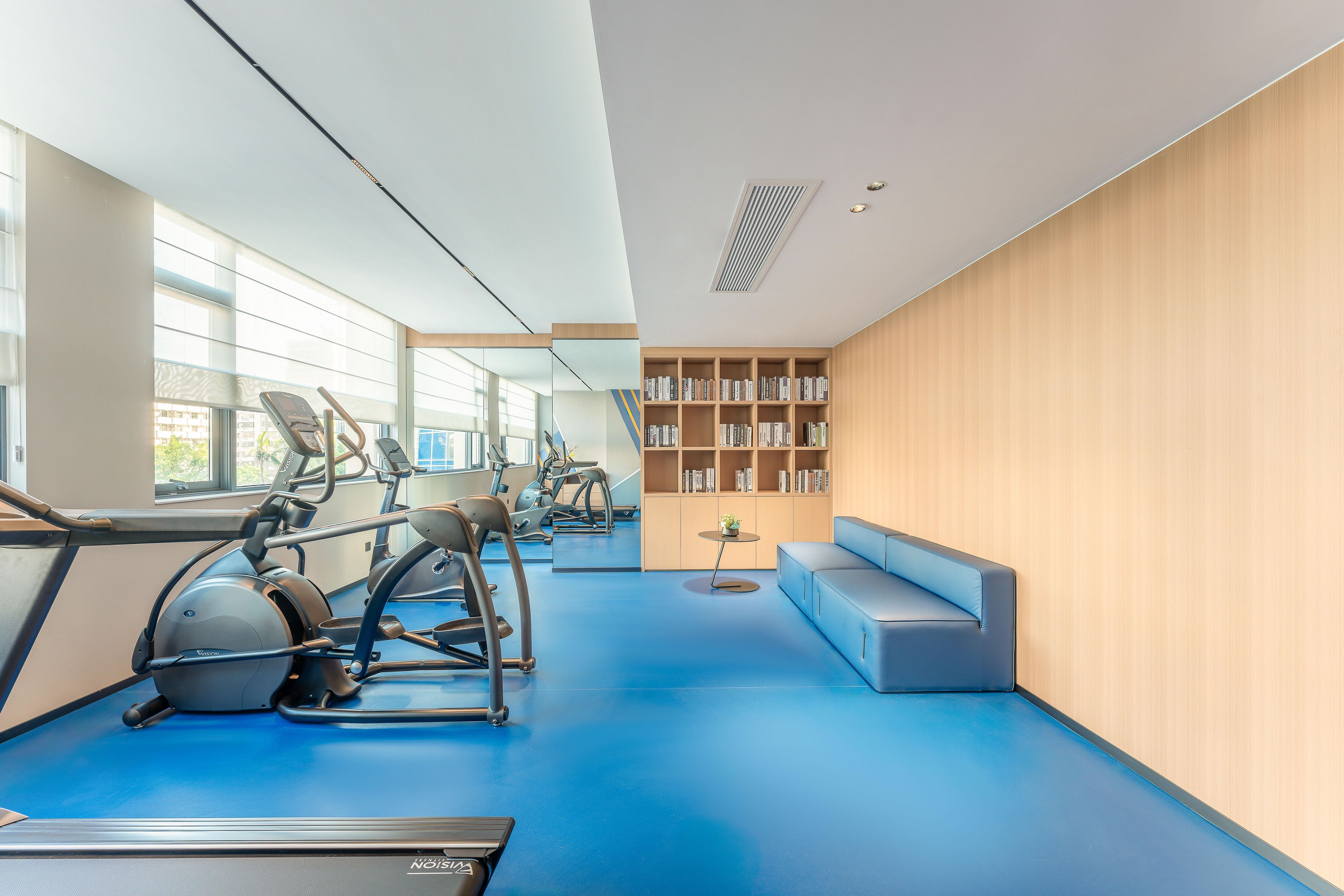 Un gimnasio moderno con equipo de ejercicio, piso azul, una estantería y un área de descanso.