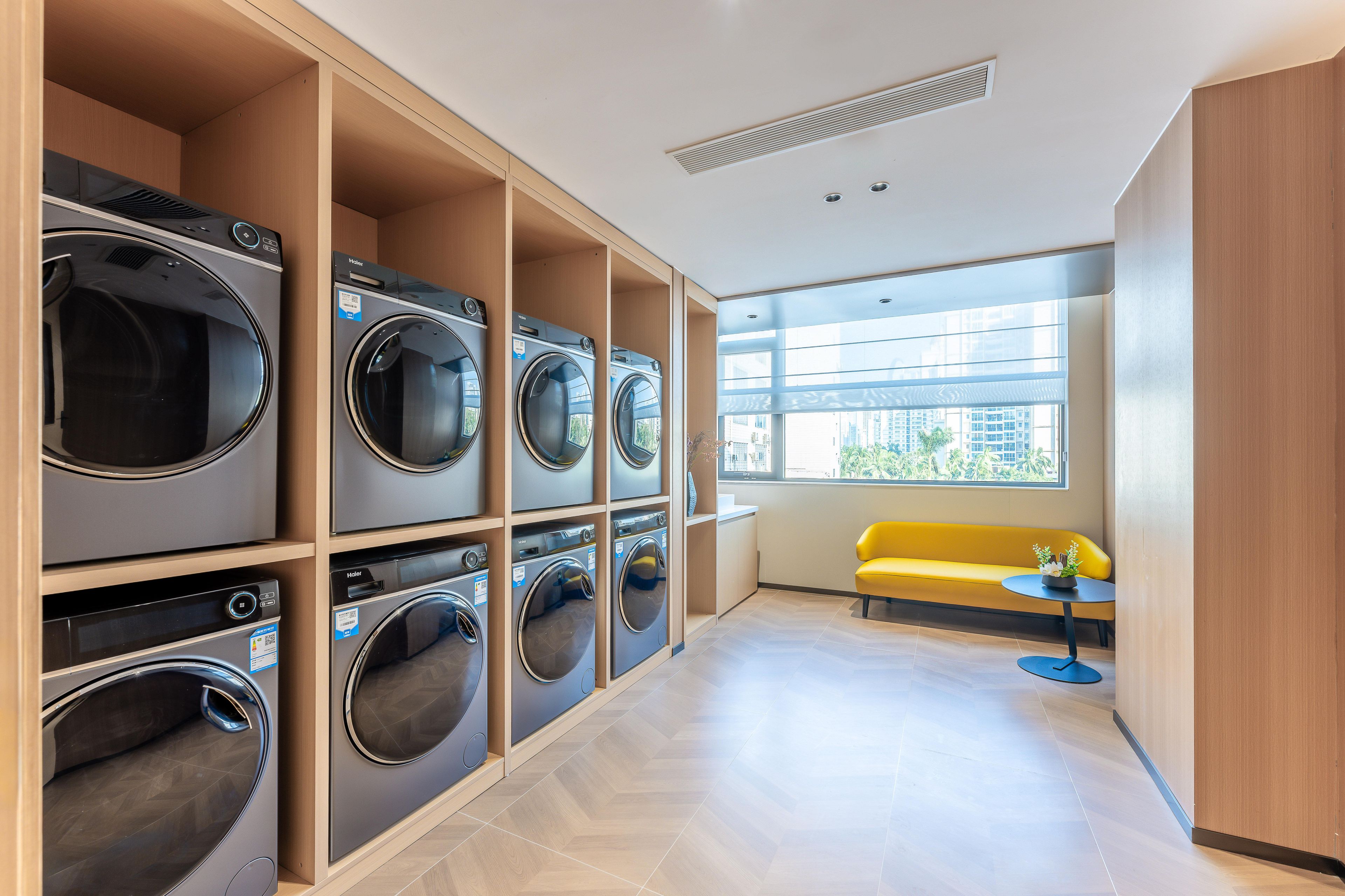 A laundry room with multiple washing machines, a yellow bench, and a small table with a plant.