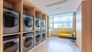 A laundry room with multiple washing machines, a yellow bench, and a small table with a plant.