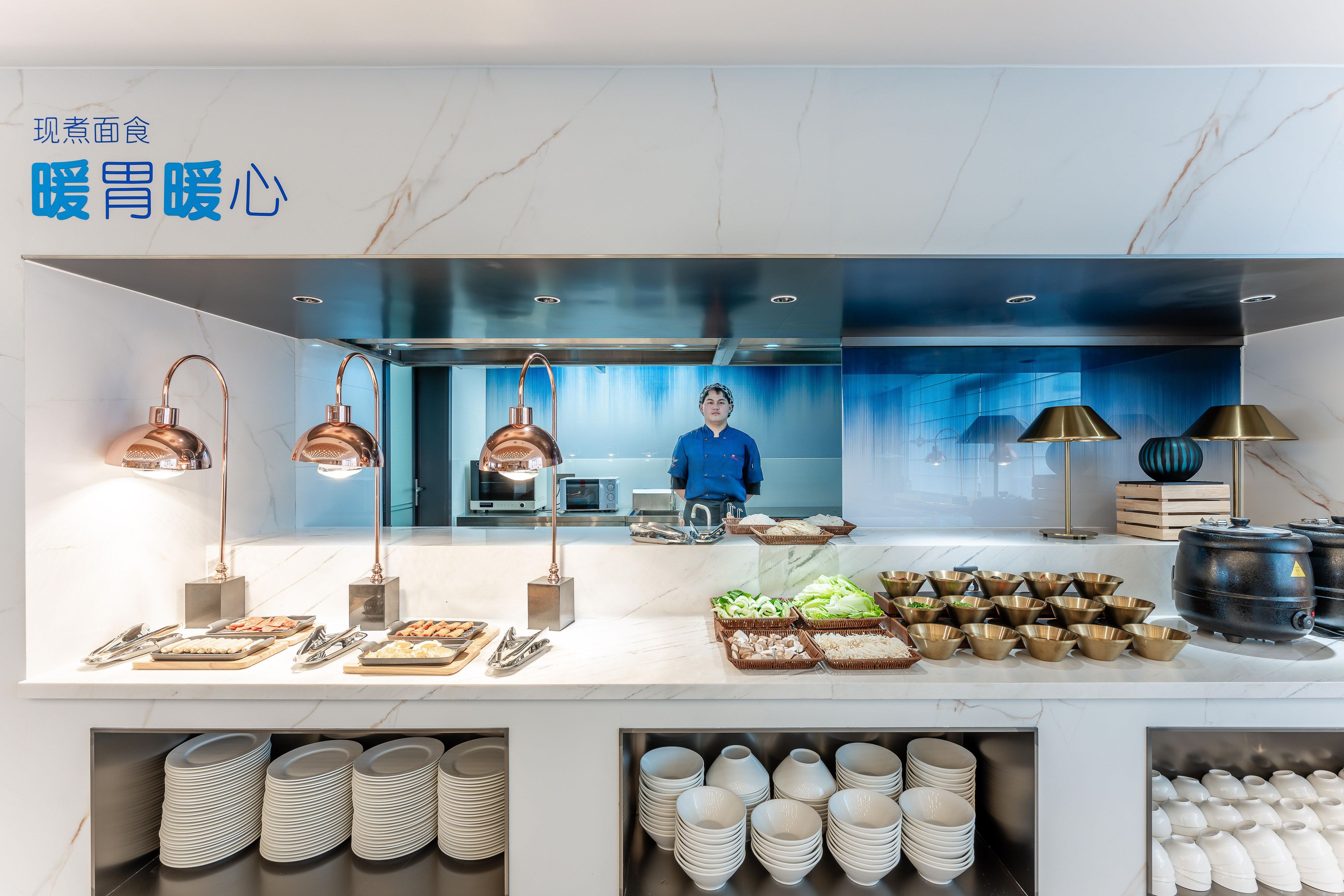 A buffet setup with various food items and utensils, a person in the background, and a modern kitchen area.
