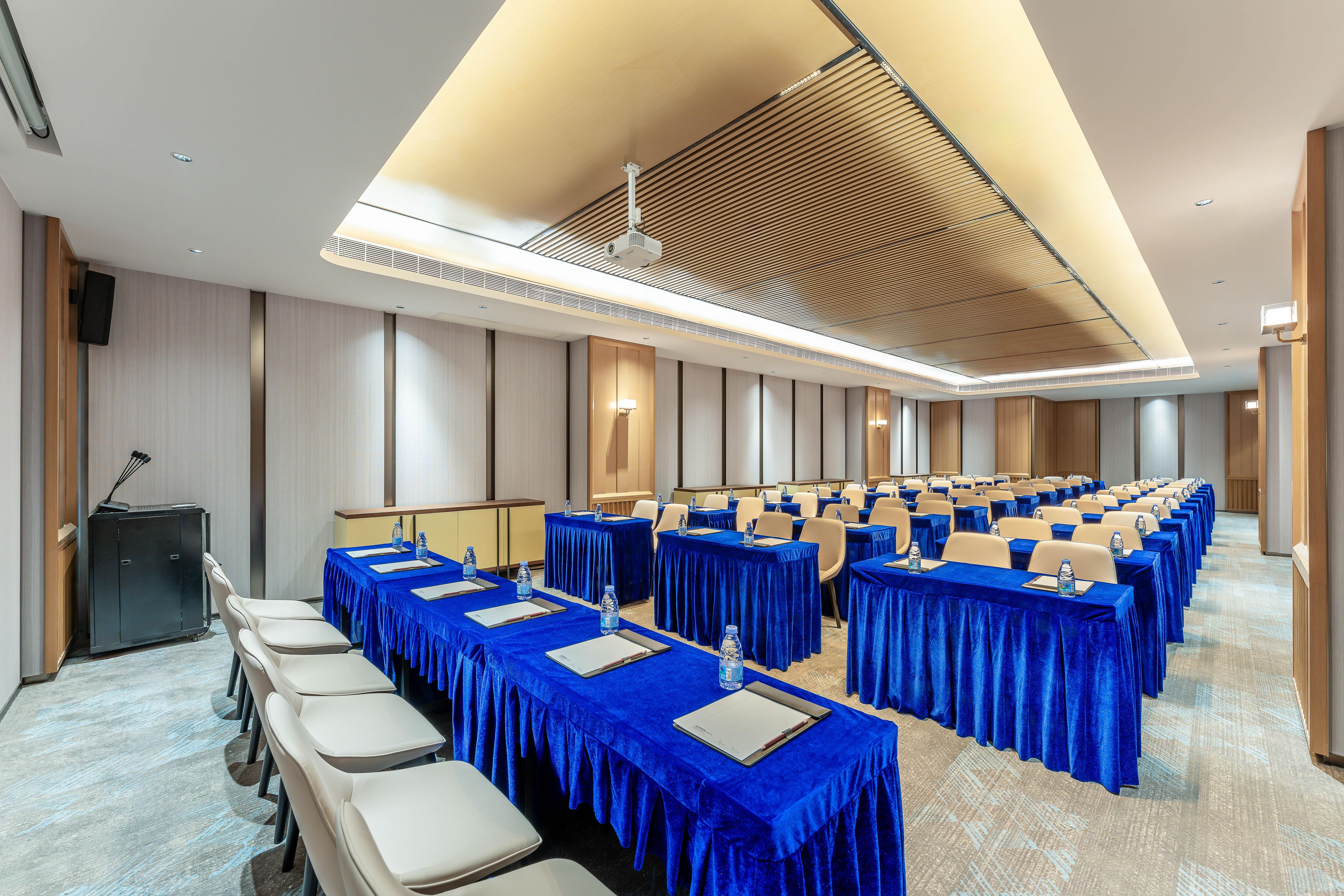 A conference room with rows of tables covered in blue tablecloths, equipped with water bottles and notepads, and a podium on the left.