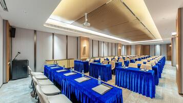A conference room with rows of tables covered in blue tablecloths, equipped with water bottles and notepads, and a podium on the left.