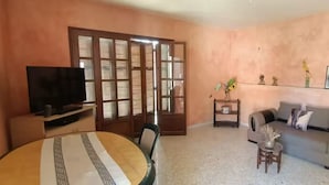 Uma sala de estar com televisão, mesa de jantar, sofá e uma estante decorativa.