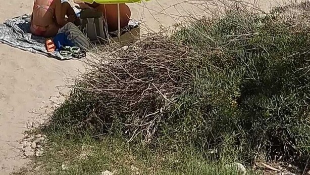 Ein Strand mit badenden und sonnenden Personen, einem grünen Sonnenschirm und felsigen Klippen im Hintergrund.