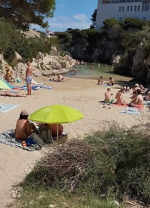 A beach with people sunbathing and swimming, a green umbrella, and rocky cliffs in the background.