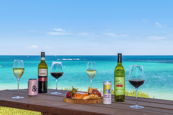 Ein Holztisch mit verschiedenen Getränken und Snacks mit Blick auf einen Strand und klares, blaues Wasser.
