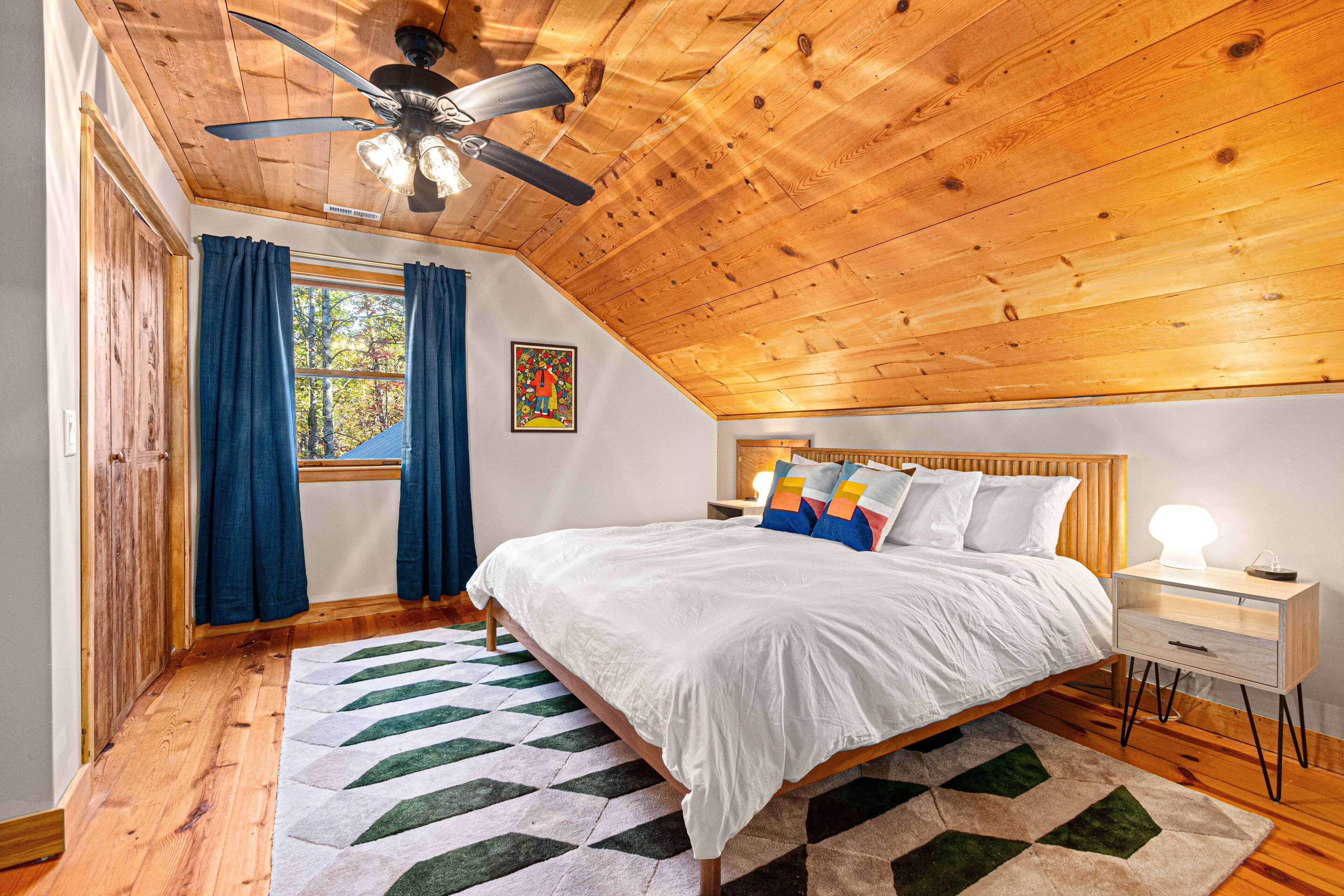 A bedroom with a wooden ceiling, a bed with white bedding, a nightstand, and a window with blue curtains.