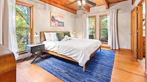 A bedroom with a wooden ceiling, a bed with white bedding, a blue rug, and large windows with curtains.